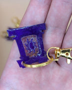 Purple keychain with gold accent held in a hand against a neutral background